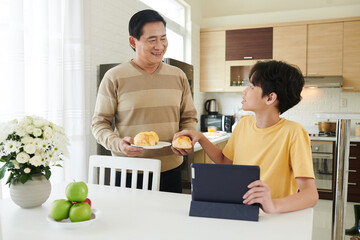 Fototapeta na wymiar Smiling father bringing plate with fresh cheese buns to his teenage son sitting at kitchen table with tablet computer and watching show