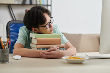 Dreaming teenage schoolboy in glasses leaning on stack of students books on his desk at home