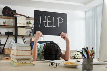 Exhausted schoolboy tired of studying sitting at desk and showing help sign