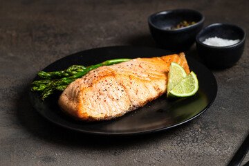 Baked salmon fillet with crispy skin,   toasted asparagus  and lime on black plate.