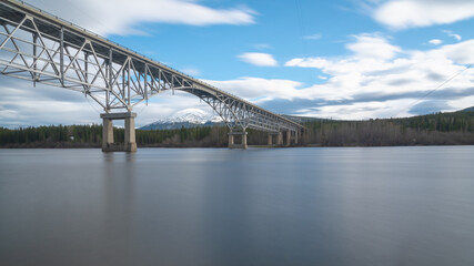 Fototapeta na wymiar Johnsons Crossing, Teslin River steel Bridge on the Alaska Highway with long exposure shot of calm, silky looking water and cloudy, blue sky sunset afternoon. 