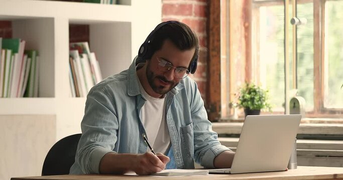Happy young 30s caucasian man in eyeglasses wearing wireless headphones, watching educational online lecture, writing notes in copybook, enjoying distant learning, getting knowledge at home office.