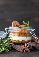 Layered dessert in glass jar with cookie crumble and whipped cream decorated with rosemary and anise, dark brown background. No bake cheesecake, trifle or pudding.