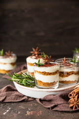 Layered dessert in glass jar with cookie crumble and whipped cream decorated with rosemary and anise, dark brown background. No bake cheesecake, trifle or pudding.