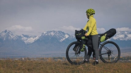 Fototapeta premium The woman travel on mixed terrain cycle touring with bikepacking. The traveler journey with bicycle bags. Sport tourism bikepacking, bike, sportswear in green black colors. Mountain snow capped.