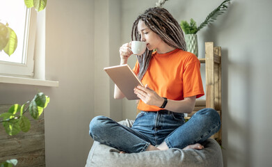 Young modern woman is sitting on a bag chair with a cup of coffee at home and using a tablet to read the news or view other information. Concept of pastime, relaxation and lifestyle