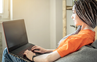 Fototapeta premium Young modern woman with dreadlocks is comfortably sitting in a chair bag at home and using a laptop for work, study or leisure. Concept of pastime, lifestyle and comfortable remotely work