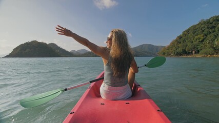 Long haired blonde woman with sunglasses rows bright pink canoe along sea bay water to beach with growing palms. Traveling to tropical countries. Sports girl is sailing on kayak in ocean.
