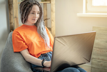 Young modern woman with dreadlocks is comfortably sitting in a chair bag at home and using a laptop for work, study or leisure. Concept of pastime, lifestyle and comfortable remotely work