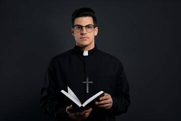 Priest in cassock with Bible on black background - obrazy, fototapety, plakaty