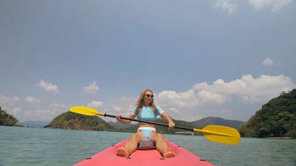 Pretty young blonde woman in stylish swimsuit and sunglasses sits on pink canoe sailing along sea...