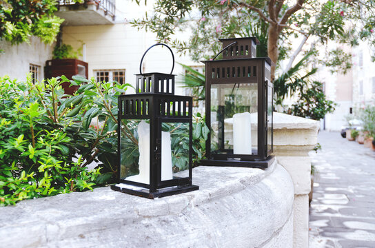 A Flashlight Candlestick, Black Vintage Decorative Lanterns With Candles Stands Outdoor In City, In Nature With Flowers And Olive Tree. Home Garden Decoration, Green Lifestyle. 