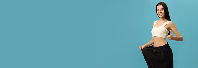 Happy young woman wearing big jeans after weight loss on turquoise background