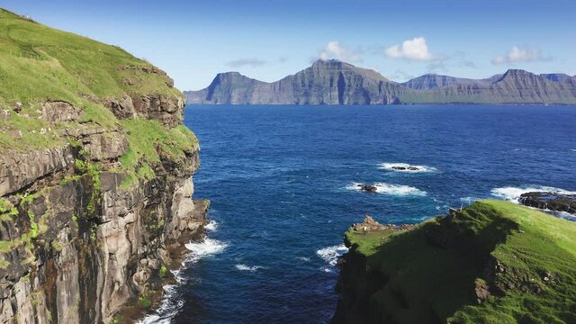 Drone flying over grass covered cliff and sea. Seagulls flying over blue rough sea near drone. In background mountain and cliff in a summer sunny day.