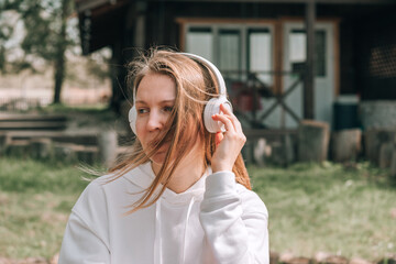 A girl in a white hoodie listens to music with headphones and relaxes sitting on a bench near a...