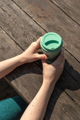 A girl holds green reuse cup in her hands against the background of a wooden table, sunlight, copy space