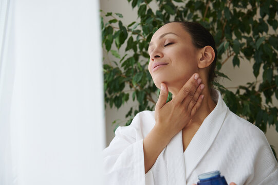 Young African American woman in white bathrobe caring of her skin, applying face moisturizer , tenderly massaging her neck