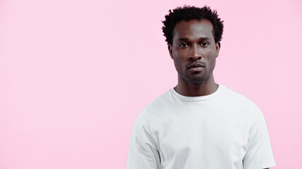 serious african american man in white t-shirt looking at camera isolated on pink