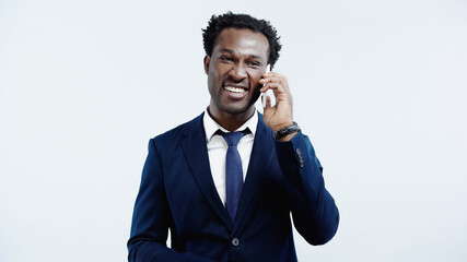 happy african american businessman in suit talking on mobile phone isolated on blue