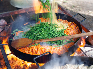 Huge pan of grilled green beans, carrot and corn cooking for catering, horizontal close-up shot of fire-fried food with wooden spoon for the restaurant on the street. 