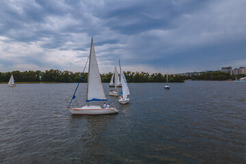 Regatta of sailing yachts with white sails on the lake in the city park. Aerial view of a sailboat in windy conditions. Summer sport yachting. Swimming in cloudy weather.