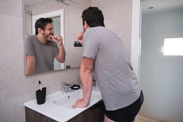 Young latin man brushing his teeth in front of the mirror in the bathroom at morning. Eco-friendly bamboo toothbrush.