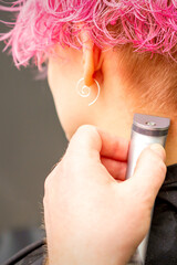 Back view of hairdresser's hand shaving nape and neck with electric trimmer of young caucasian woman with short pink hair in beauty salon