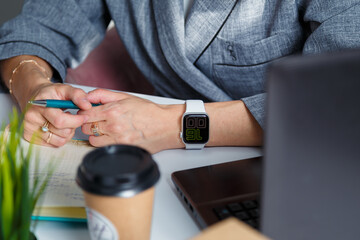 Business woman in grey jacket holds pen. Hands with watch on table. Laptop, potflower, coffee cup on foreground. Office work