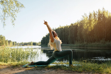 Yoga in nature. Young woman practicing yoga on lake at sunset, summer. Fitness model standing on sports mat in anjaneyasana pose, performing yoga asana, outdoors. Active healthy lifestyle concept