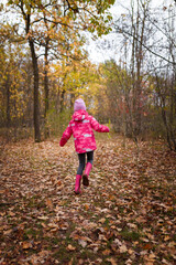 Little girl in pink jacket, knit cap and resin boots happily running in autumn forest on golden leaves cover backwards to camera