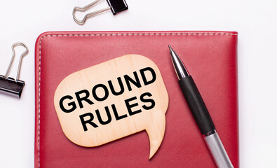 On a light background there are black paper clips, a pen, a burgundy notepad a wooden board with the text GROUND RULES.