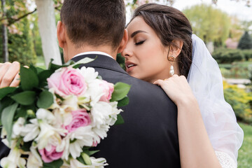Obraz na płótnie Canvas Pretty bride with blurred bouquet hugging groom in park