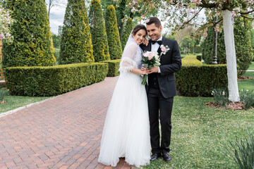 Cheerful bride with bouquet hugging groom in park