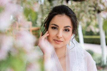 Fototapeta premium Pretty bride in white veil looking at camera in park