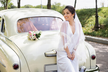 Happy bride in wedding dress standing near bouquet on retro auto