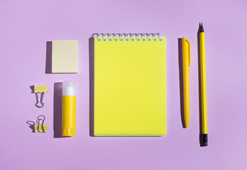 Yellow stationery supplies on bright purple background. Notepad with empty blank, stickers, glue, pen and pencil. Back to school concept. Top view, flat lay, mockup