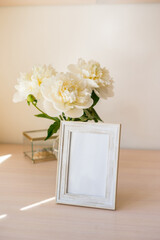 Portrait white picture frame mockup on wooden table. Modern glass vase with peony. White wall background. Scandinavian interior. Vertical.