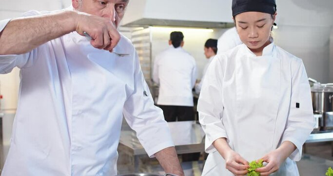 Close up of old Caucasian male chef tasting prepared dish talking to young Asian female professional cook washing salad. Backstage workflow in kitchen, restaurant, team cooking dishes with vegetables