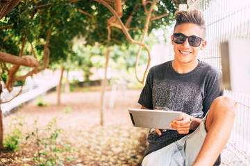 Young boy teenager age use tablet device outdoor in the park smiling and enjoying connection technology - happy youth people man portrait in nature garden - Powered by Adobe