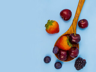 summer concept scattering of juicy berries of strawberries, blackberries, blueberries and cherries in a wooden spoon on a clean bright blue background, fresh idea for design