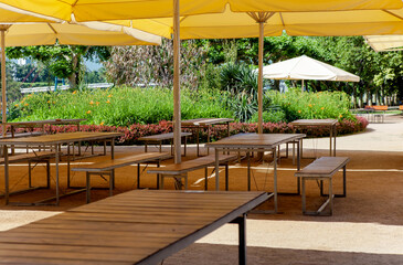 Outdoor restaurant in Germany located on public park, among greenery. Enclosed beer garden with empty tables, chairs under yellow umbrellas. Corona crisis empty beer garden without guests