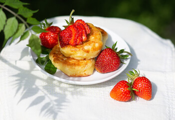 Beautiful summer breakfast - cheesecakes pancakes with fresh strawberries on a white plate. Tea in a tnka porcelain cup, sun glare and shadows. Linen tablecloth, rustic style, provence