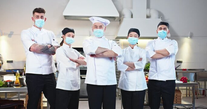 Team Of Multi-ethnic Professional Diverse Male And Female Chefs In Medical Masks Standing At Restaurant Kitchen And Looking At Camera Before Working Day During Covid Pandemic. Cookery, Job Concept