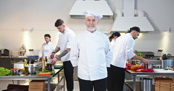 Old Caucasian senior male chef in hat standing at famous restaurant kitchen looking at camera while his team of young professional male and female cooks working preparing meals, culinary job