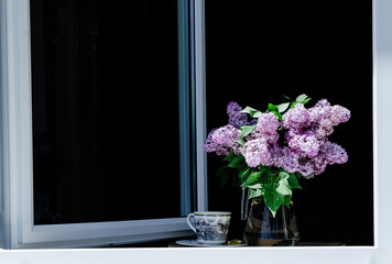 A lilac flowers bouquet and a cup of tea standing on a window sill.