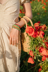 Fototapeta na wymiar Summer flowers in straw wicker bag basket. Woman hand with accessory made of colored beads close up holding bouquet of red flowers. Rural country style romance. Vertical composition