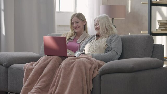 Wide shot portrait of two laughing senior Caucasian women surfing social media on laptop talking. Happy cheerful beautiful old ladies enjoying meeting in living room indoors gossiping. Friendship