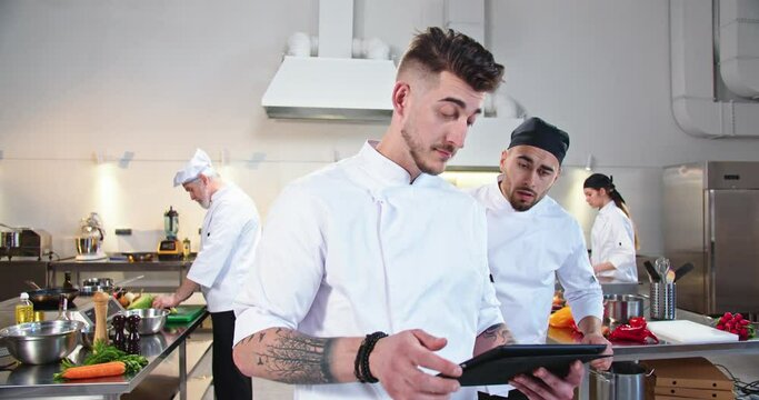 Portrait of young handsome Caucasian male chef standing in restaurant kitchen typing on tablet device and talking to busy guy cook worker showing order meal. Workflow process, team preparing food