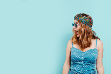 Young blonde girl with a bright headscarf looking surprised to the side with her mouth open and a pair of sunglasses with a denim top on a blue background. summer concept. copy space