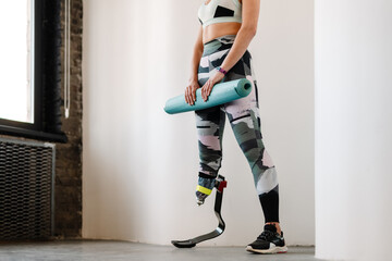 Young sportswoman with prosthesis posing with fitness mat indoors